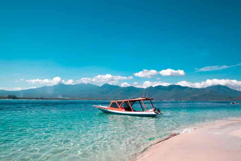 Small red-roofed boat floating on clear turquoise water near a white-sand beach at the Gili Islands, Lombok, Indonesia.