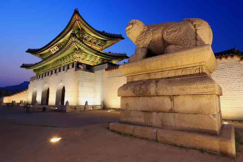 Illuminated Gyeongbokgung Palace main gate with stone guardian statue at dusk in Seoul, South Korea.