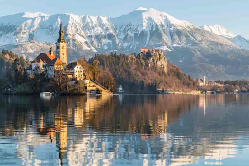 Lake Bled with Bled Island and Church of the Assumption reflected in the water, surrounded by snowy Julian Alps in Slovenia.