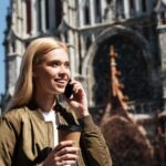 Smiling traveler using her phone overseas, holding a takeaway coffee in front of a historic European cathedral