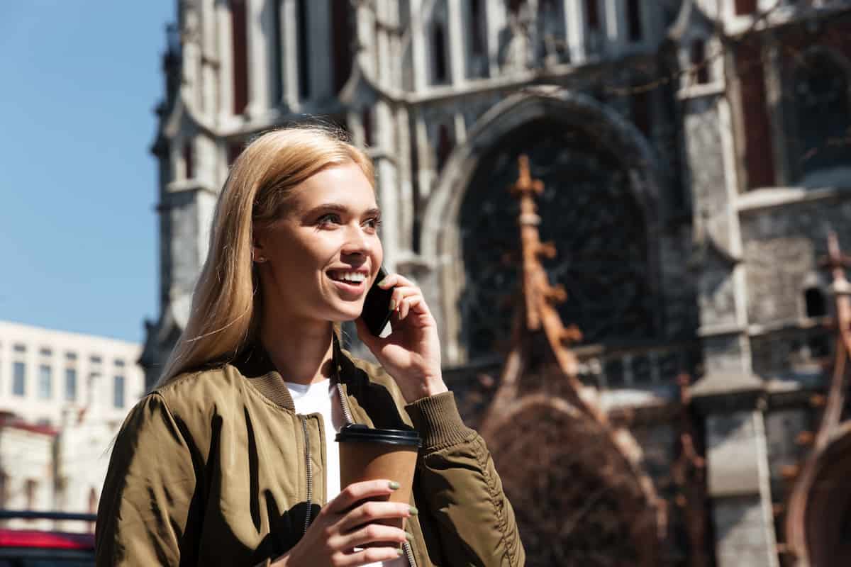 Smiling traveler using her phone overseas, holding a takeaway coffee in front of a historic European cathedral