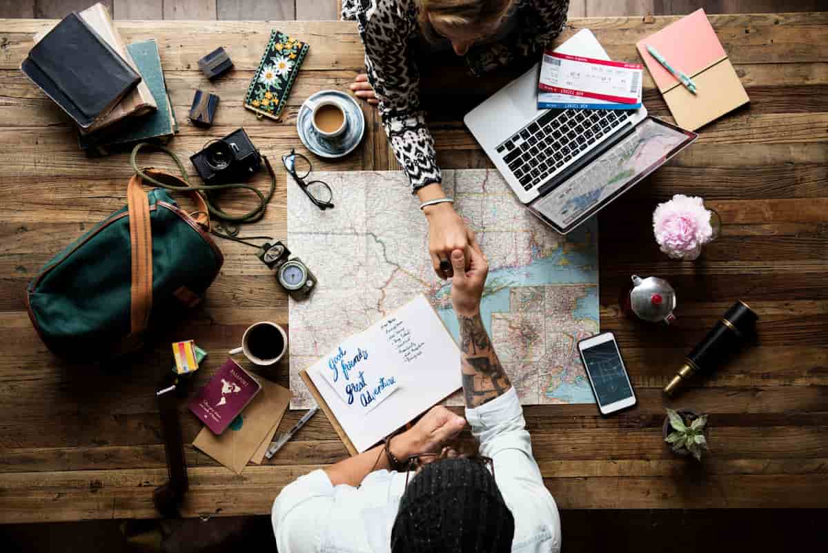 Overhead view of two travelers planning a trip with a map, laptop, passports, tickets and coffee on a wooden table.