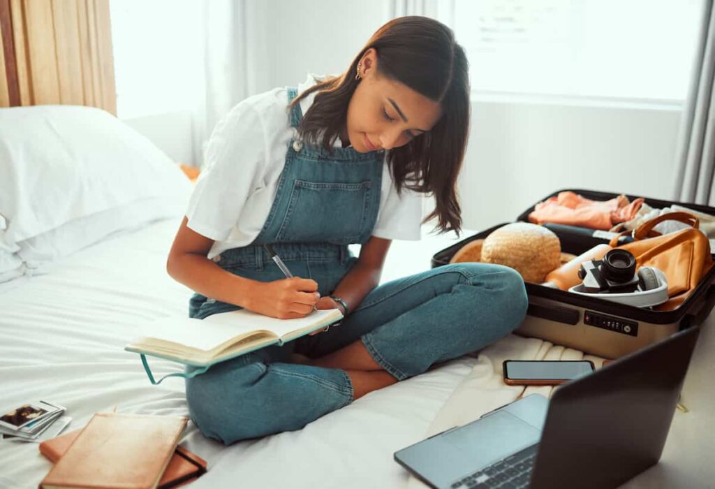 Traveler sitting on a bed writing a packing checklist beside an open suitcase, laptop and phone.