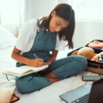 Traveler sitting on a bed writing a packing checklist beside an open suitcase, laptop and phone.
