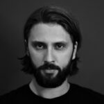 Black and white portrait of a bearded man with medium-length hair, looking directly at the camera with a serious expression.