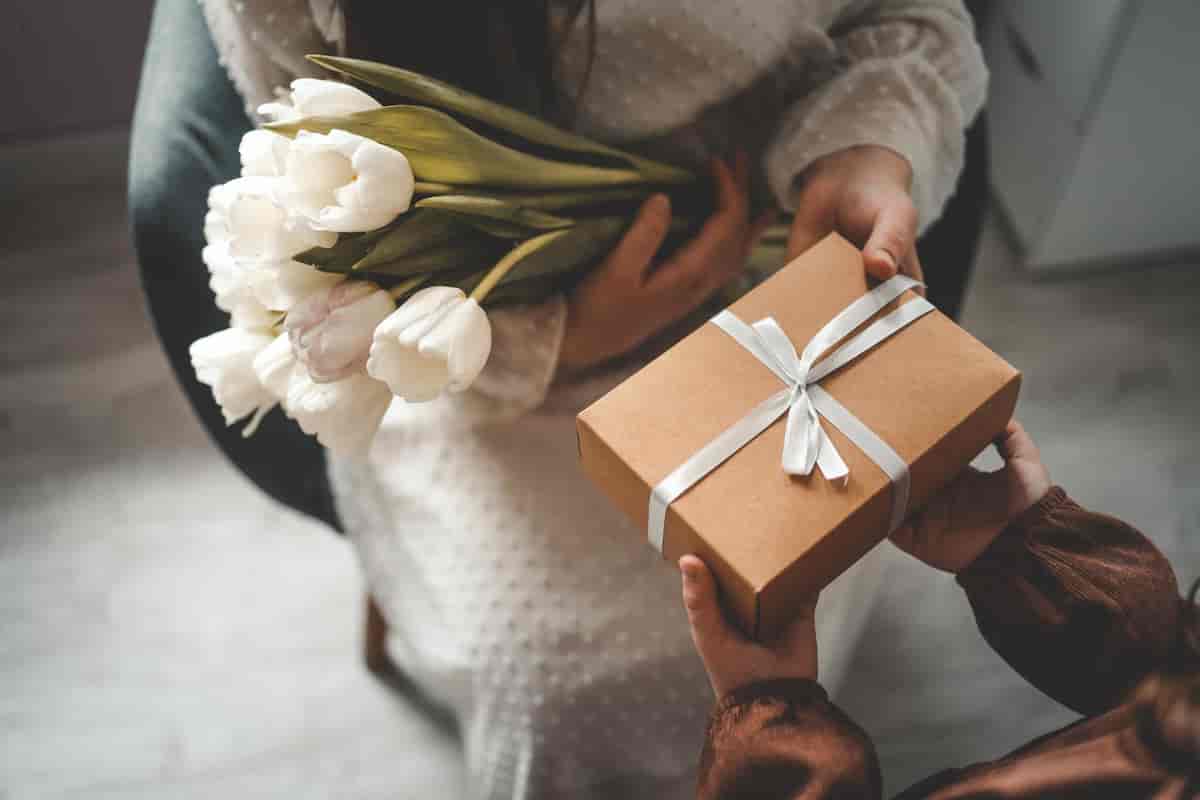 Person holding a bouquet of white tulips receiving a beautifully wrapped gift box tied with a white ribbon