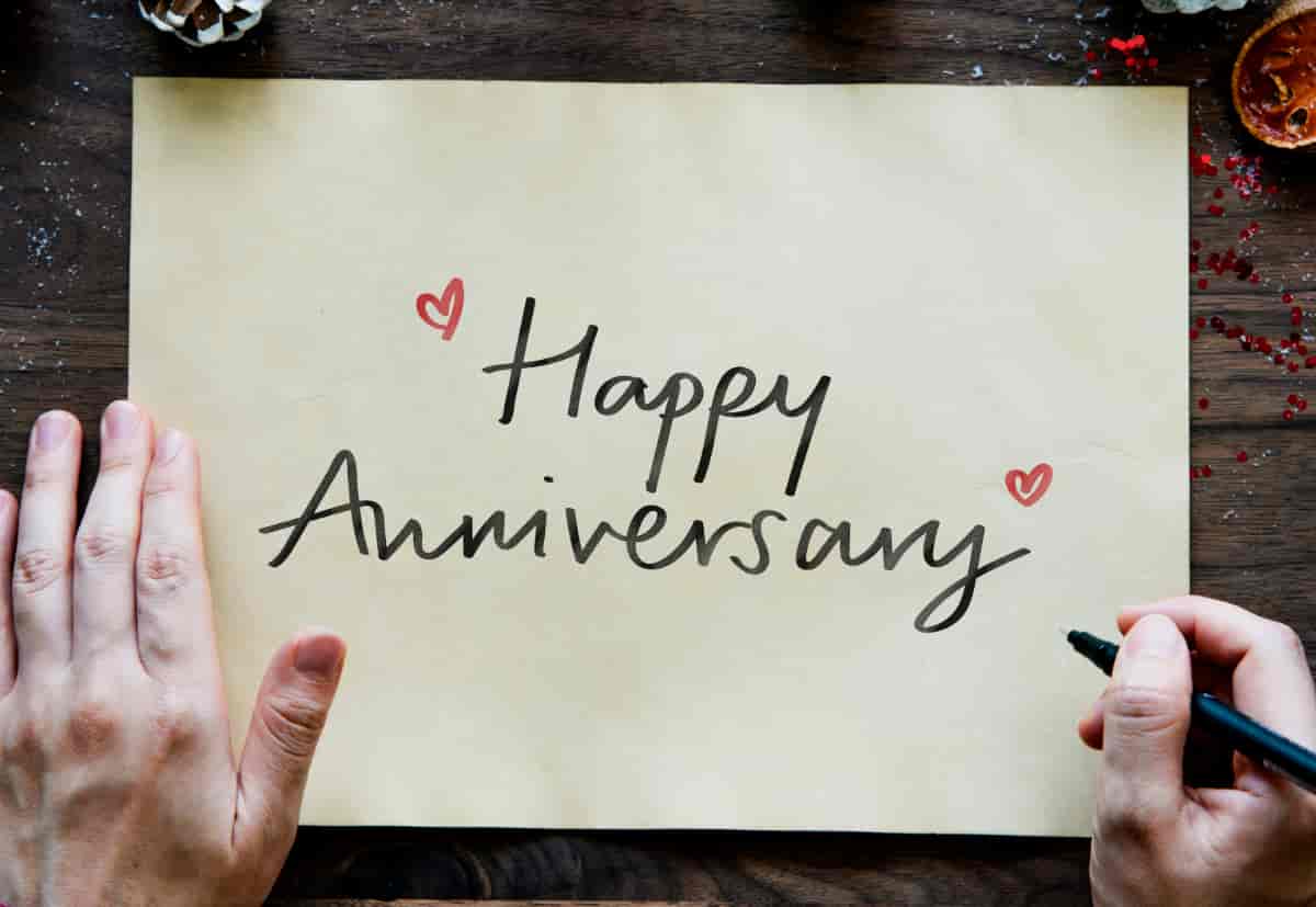 Handwritten happy anniversary message with red hearts on beige paper, held by two hands with a black marker on a wooden surface